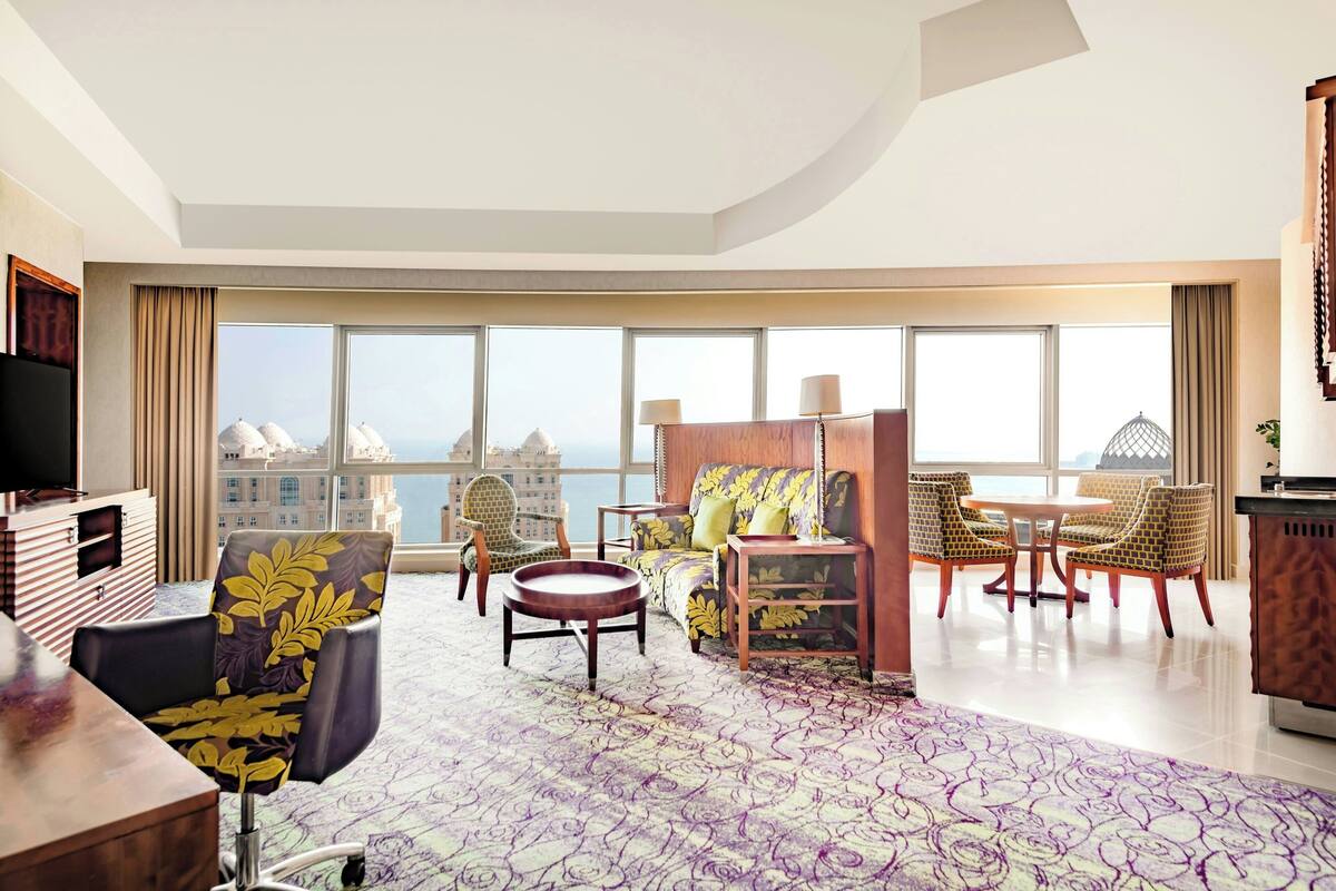 Habitación de hotel moderna con un amplio ventanal que ofrece vistas a la ciudad, una zona de estar acogedora con sofá y sillones, un televisor de pantalla plana y una mesita de madera.