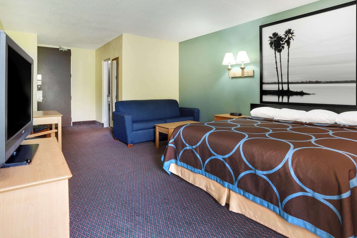 A hotel room with a bed, a TV, a sofa, a desk, and a wall with a painting.