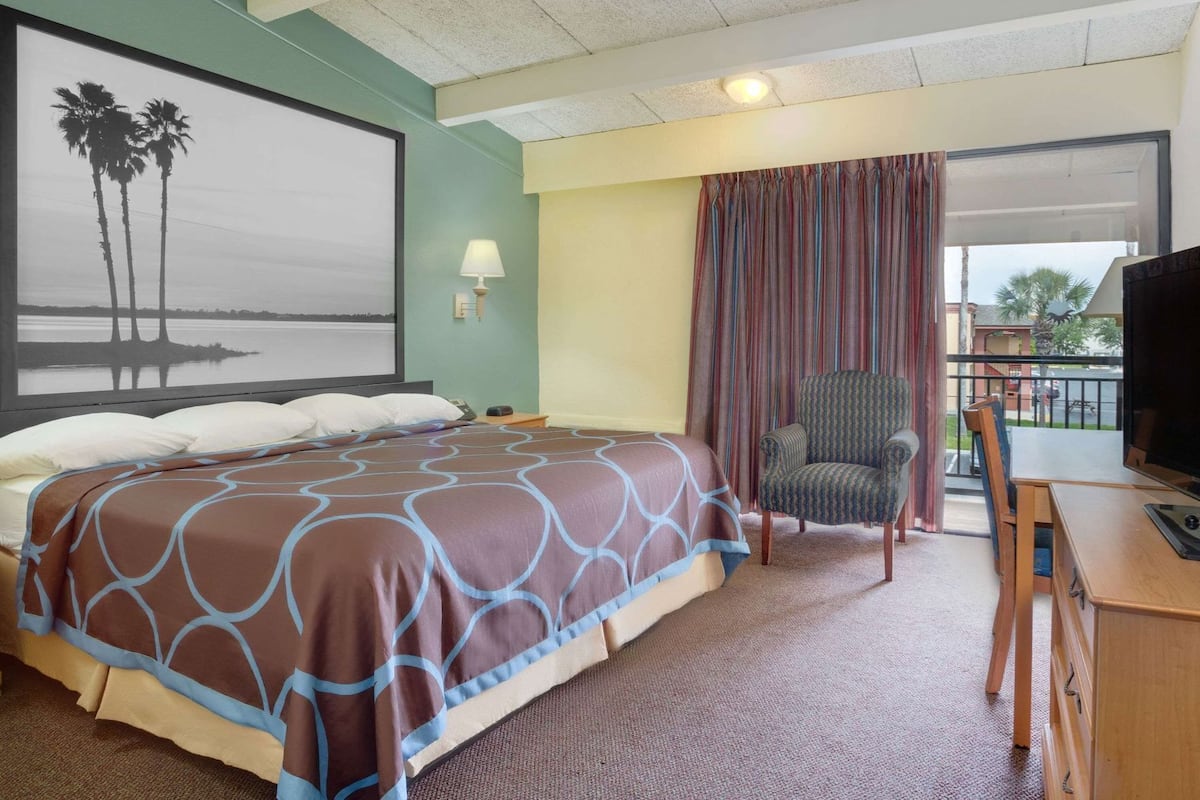 A hotel room with a bed, a chair, a desk with a television, a window with curtains, and a painting on the wall.