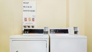 Two front-loading washing machines with control panels, a sign above them listing available detergents, and a wall-mounted electrical outlet. - Super 8 by Wyndham St. Augustine (St. Augustine)