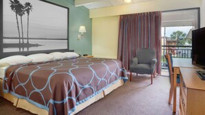 A hotel room with a bed, a desk, a chair, a TV, a window with curtains, and a painting of palm trees. - Super 8 by Wyndham St. Augustine (St. Augustine)