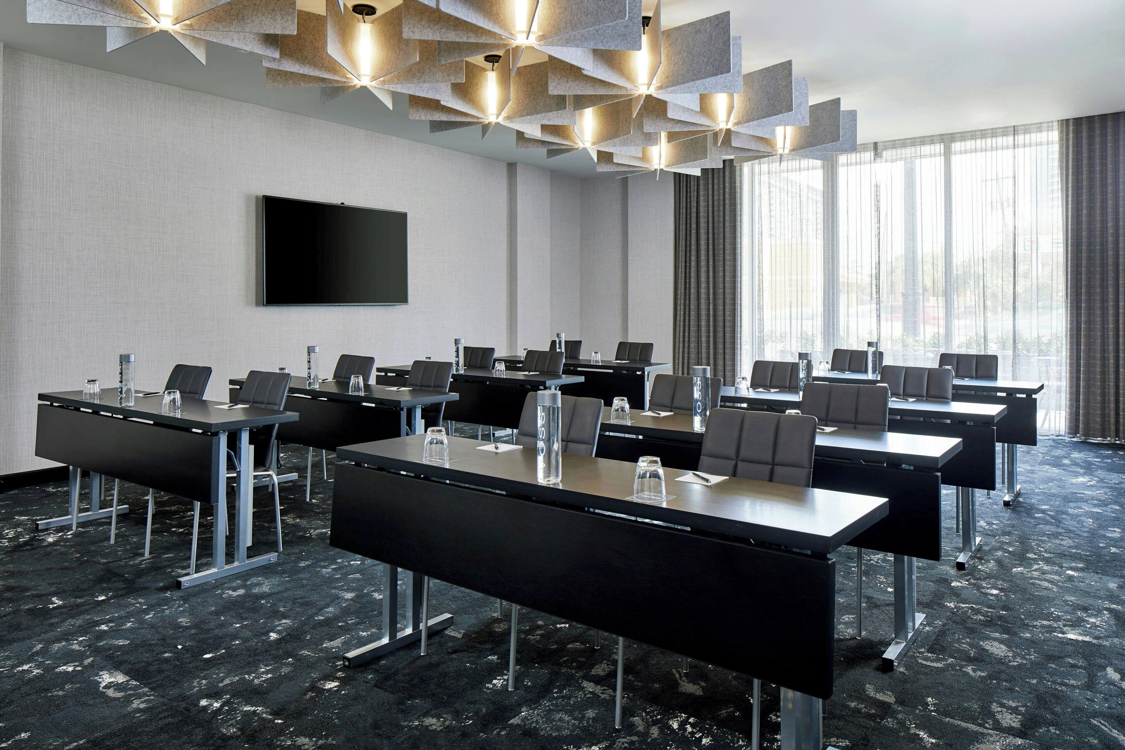 Une salle de conférence équipée de plusieurs tables, de chaises, d’un téléviseur à écran plat et d’une grande fenêtre avec des rideaux.