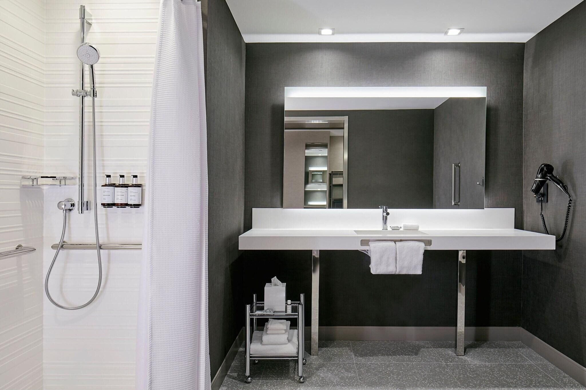 Un baño moderno con un espejo grande, un lavamanos blanco y una ducha con cabezal de ducha de mano.