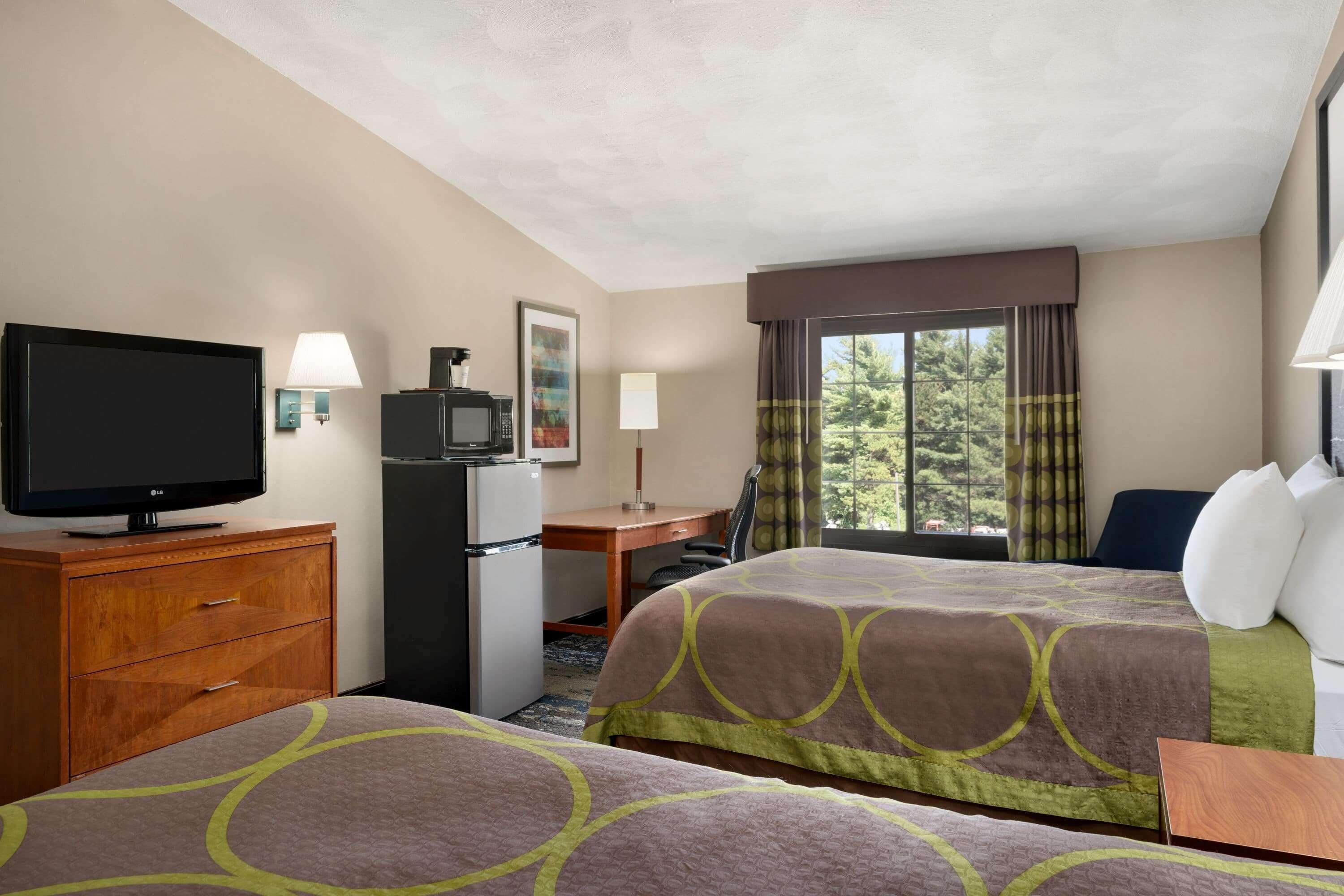 A hotel room with two beds, a desk, a television, a refrigerator, and a window with curtains.