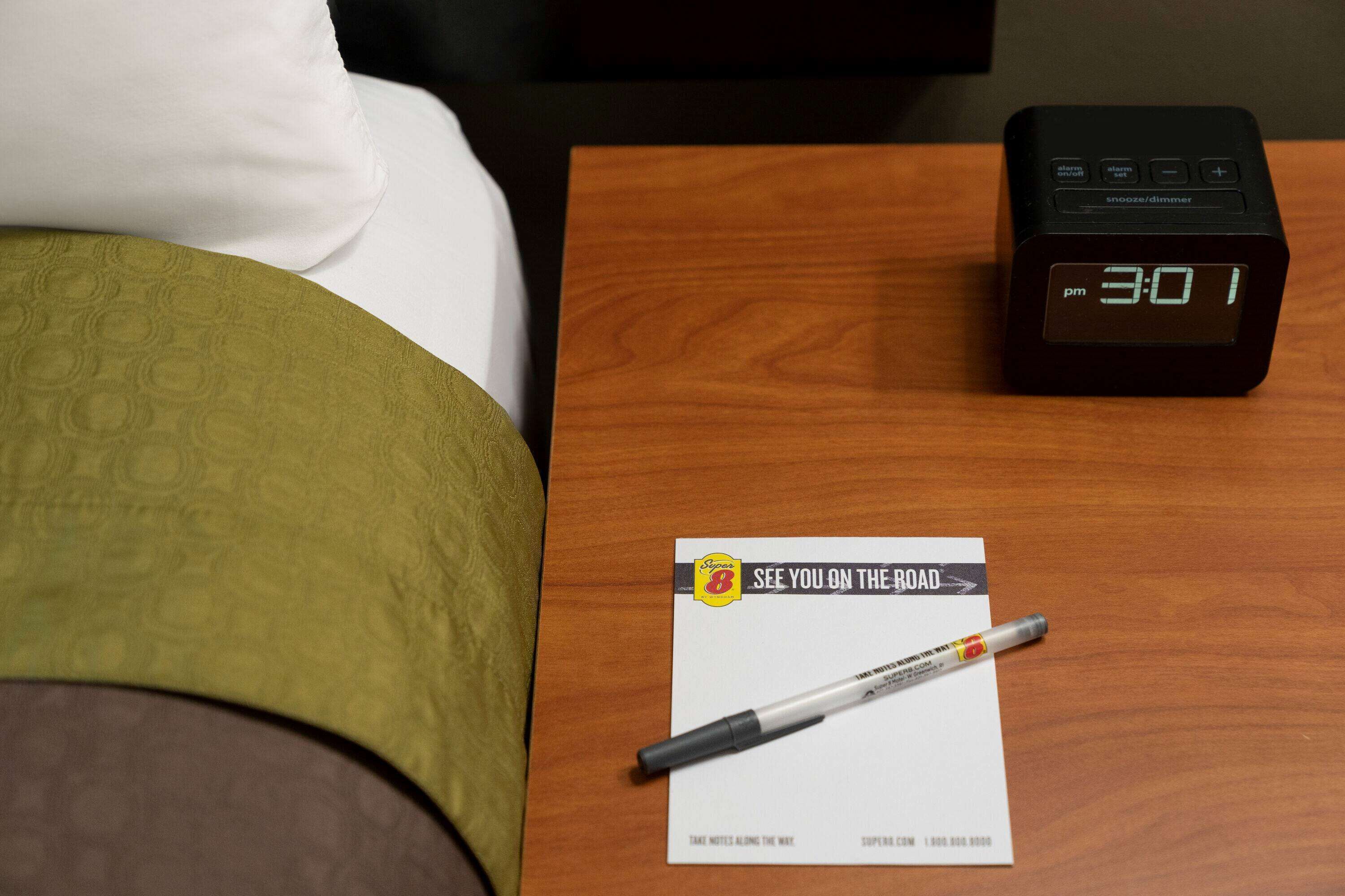 A wooden nightstand with a digital clock displaying 3:01 PM, a pen, and a business card on a hotel bed.