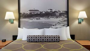 A hotel room with a bed, two bedside lamps, a framed photograph of a coastal scene, and a patterned pillow.