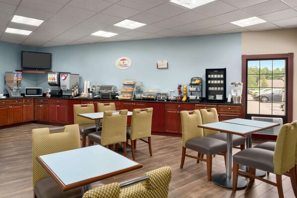 Uma área de café da manhã com mesas, cadeiras, uma máquina de café e uma televisão na parede.