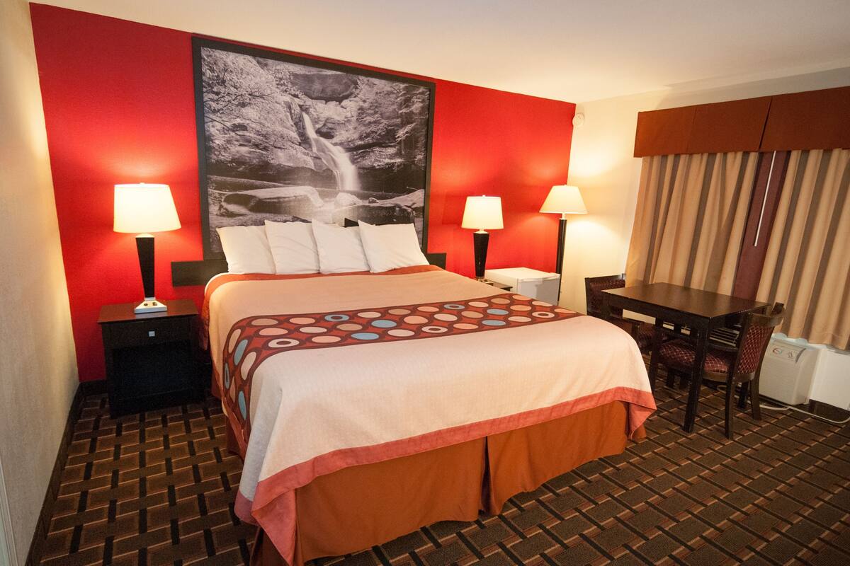 A hotel room with a bed, two bedside tables with lamps, a desk with chairs, and a large framed picture on the wall.