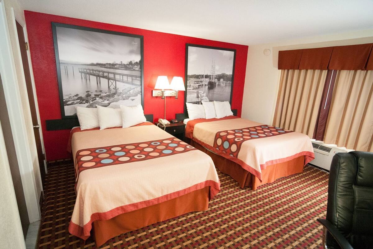A hotel room with two beds, a red wall, a patterned carpet, and a large window with curtains.