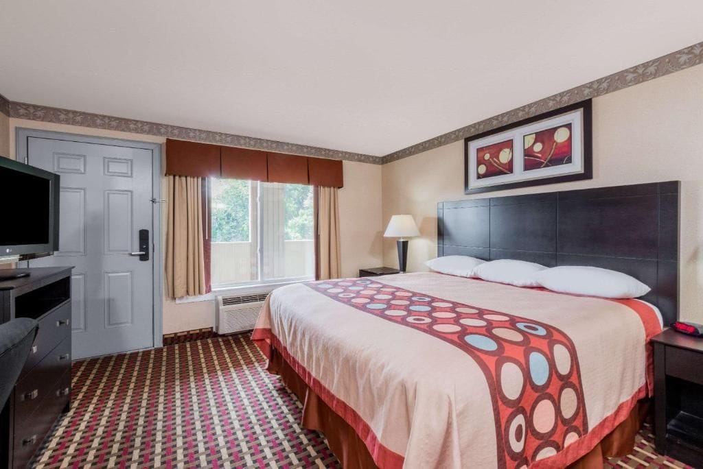 Una habitación de hotel con cama, escritorio, televisor, ventana con cortinas y una obra de arte en la pared.