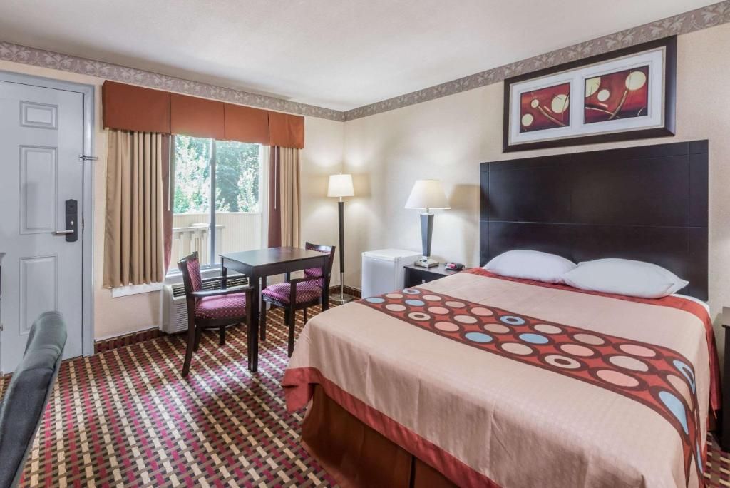 A hotel room with a bed, a desk, a chair, a window with curtains, a lamp, and artwork on the wall.