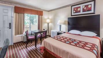 A hotel room with a bed, a desk, a chair, a window with curtains, a lamp, and artwork on the wall.