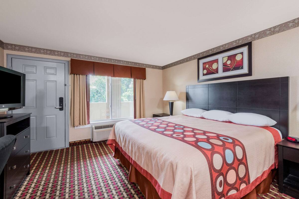 A hotel room with a bed, a desk, a television, a window with curtains, and a patterned floor.