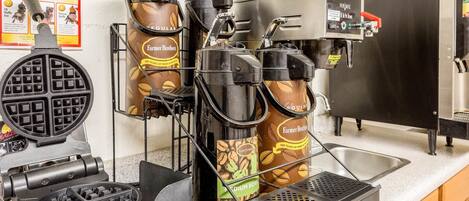Una estación de café con una máquina para gofres, dos cafeteras y un lavamanos.