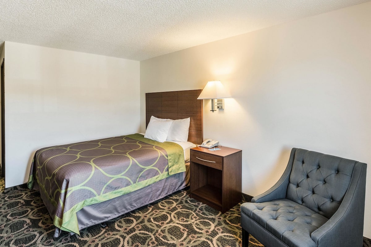 A hotel room with a bed, a nightstand, a chair, and a wall-mounted lamp.