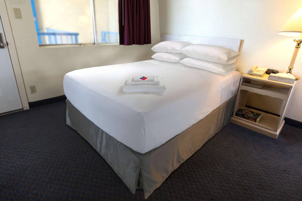 A hotel room with a neatly made bed, a bedside table with a lamp, and a small shelf with a book.