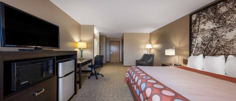 A hotel room with a bed, a desk with a computer, a television, a refrigerator, and a lamp.