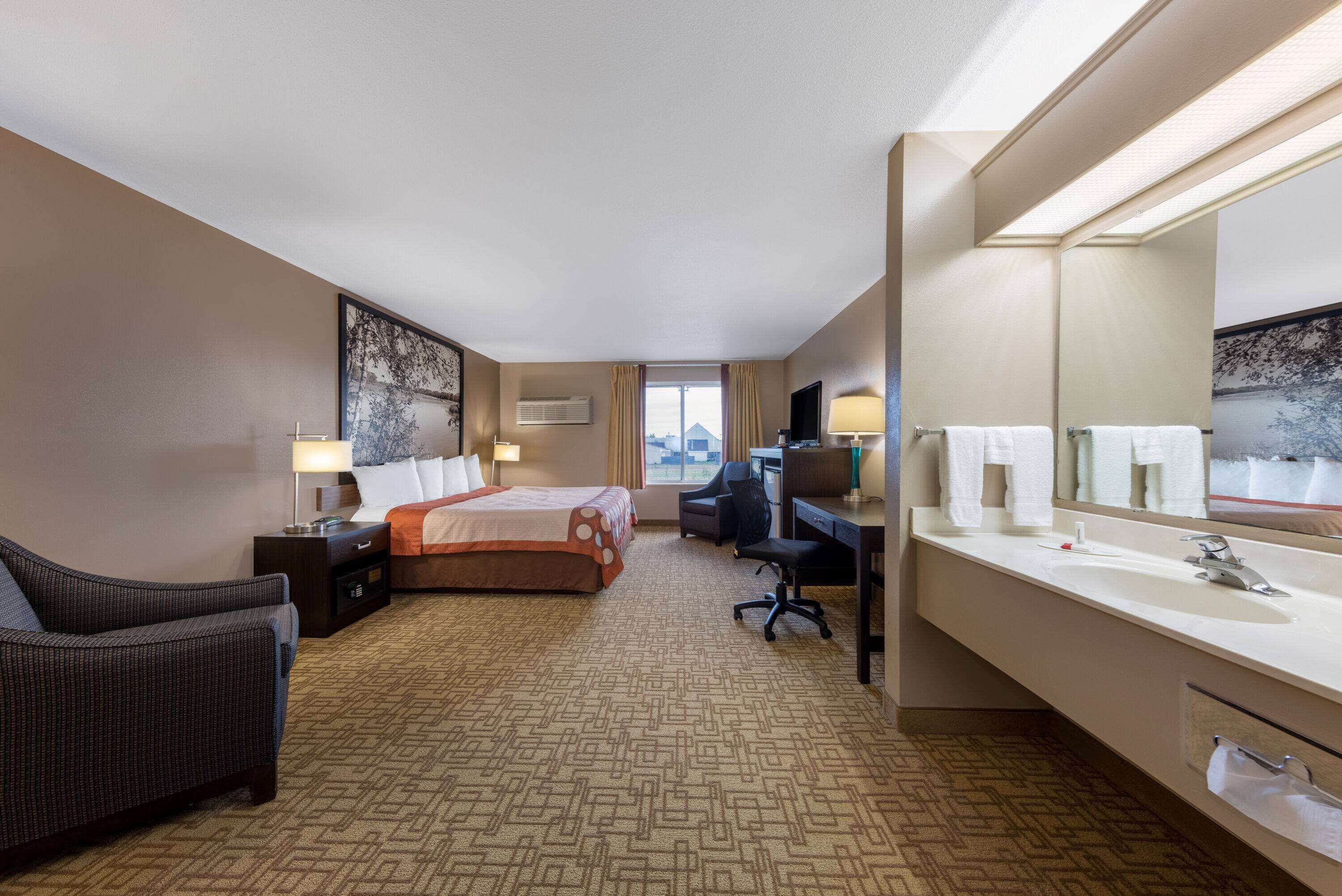 A hotel room with a bed, a desk, a chair, a large mirror, and a bathroom with a sink and toilet.