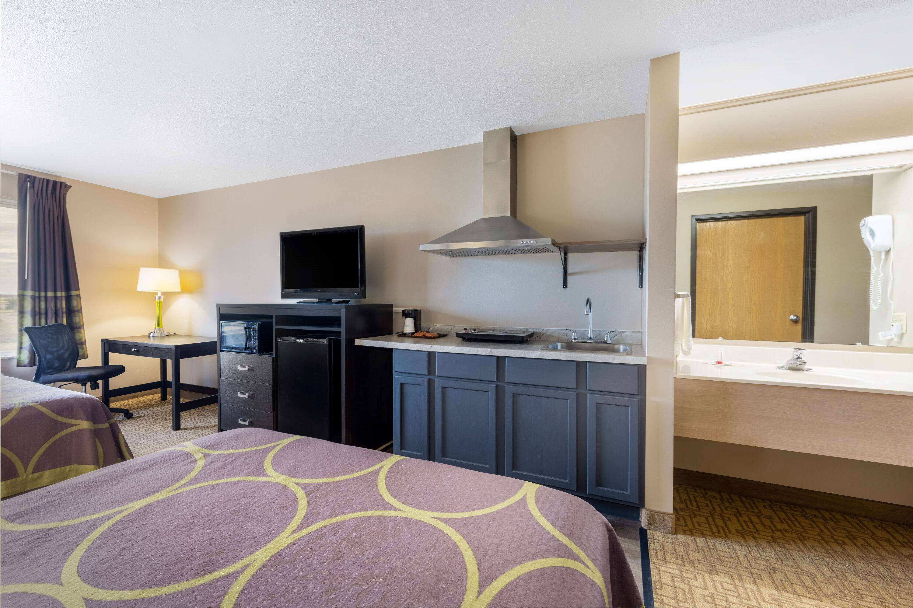 A hotel room with a bed, desk, kitchenette, and a bathroom with a mirror and sink.