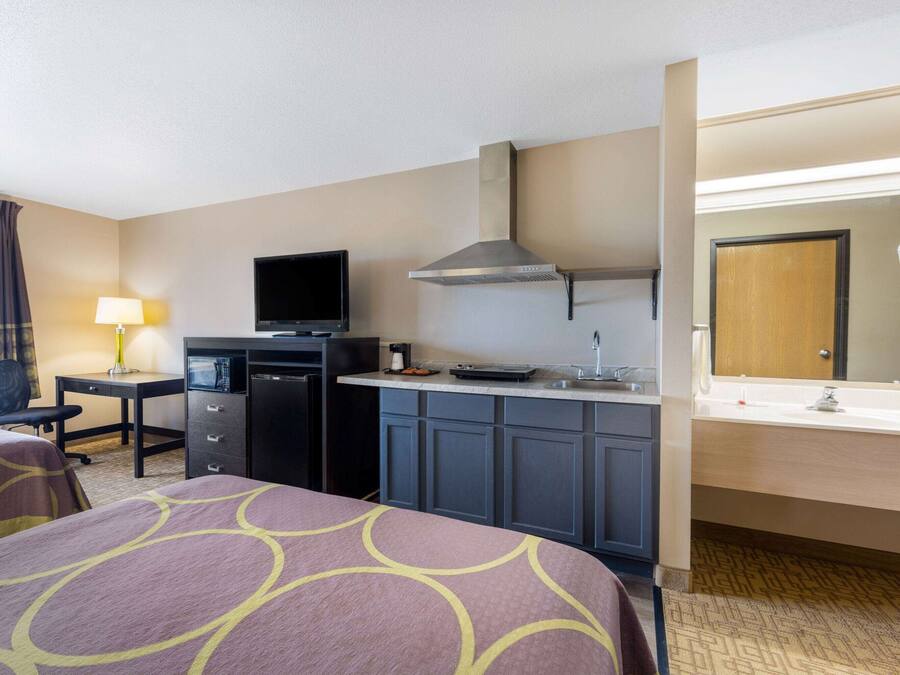 A hotel room with a bed, desk, kitchenette, and a bathroom with a mirror and sink.