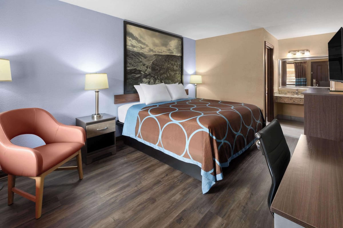 Habitación de hotel con cama, escritorio, silla, mesita de noche, lámpara y un cuadro en la pared.