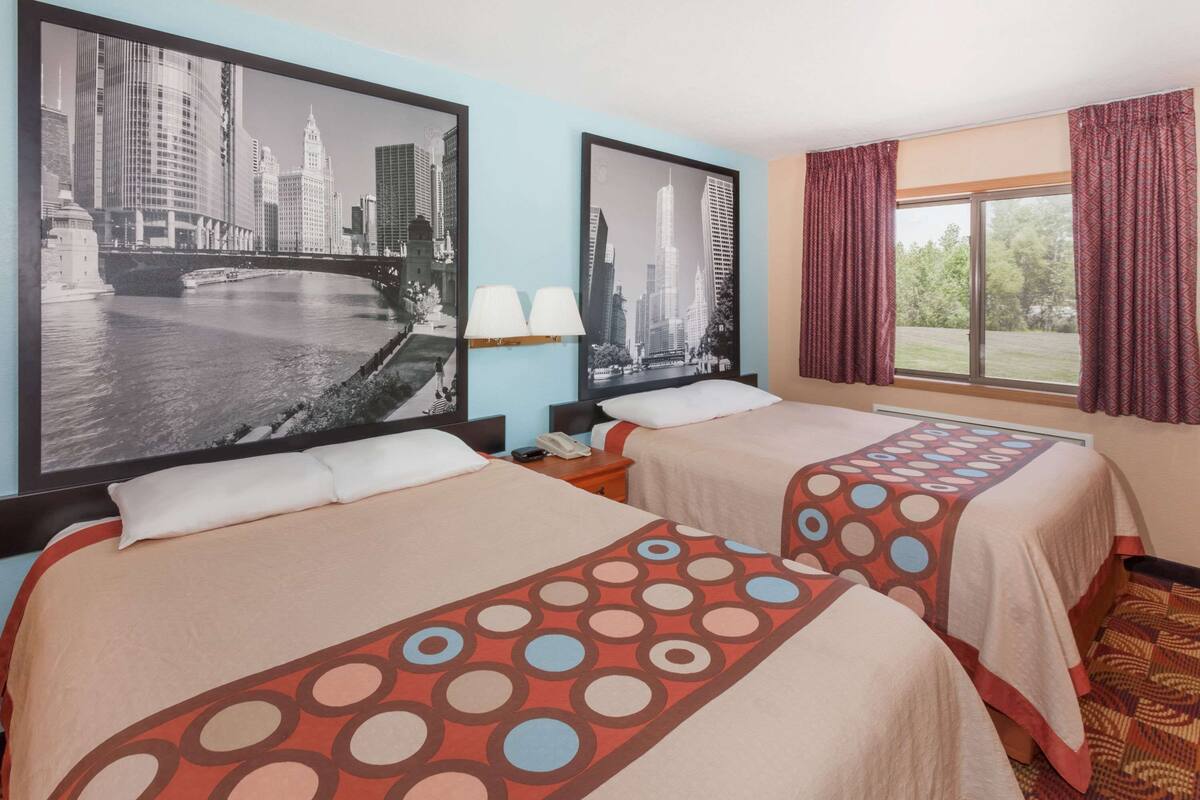 Habitación de hotel con dos camas, un cuadro de paisaje urbano en la pared, una ventana con cortinas y una mesita de noche con lámpara.