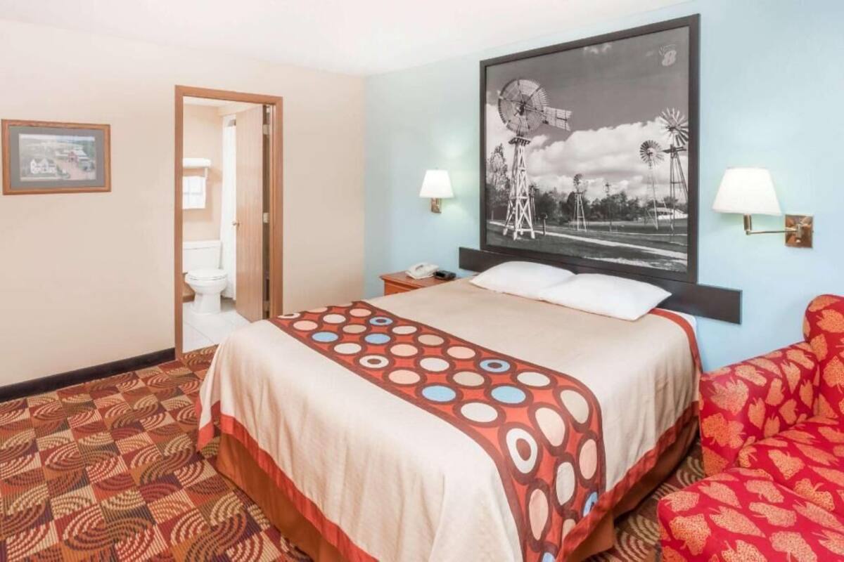 Una habitación de hotel con una cama, un sofá rojo estampado, una lámpara fijada a la pared, un cuadro enmarcado y una pintura grande de un molino.