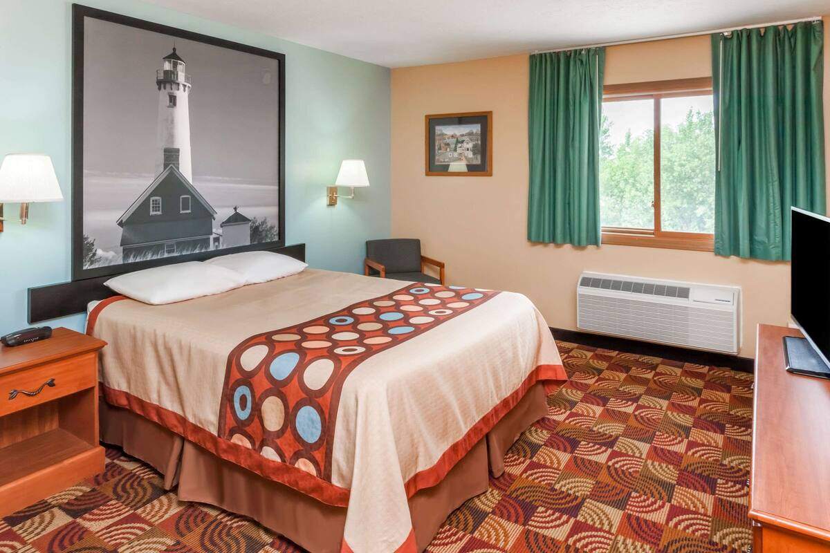 Habitación de hotel con cama, escritorio, silla, televisor, ventana con cortinas, un cuadro en la pared y unidad de aire acondicionado.