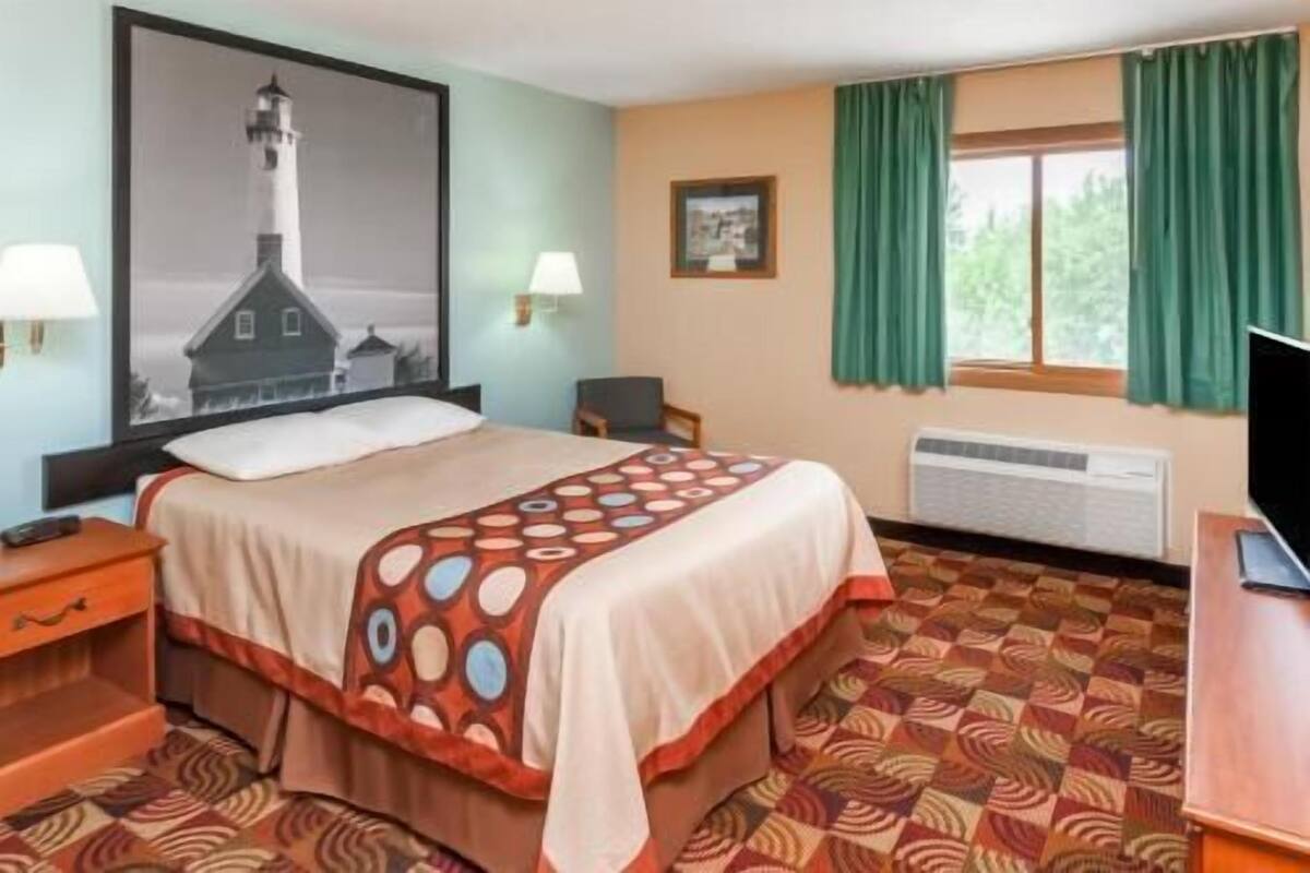 Habitación de hotel con cama, escritorio, silla, un cuadro en la pared y ventana con cortinas.