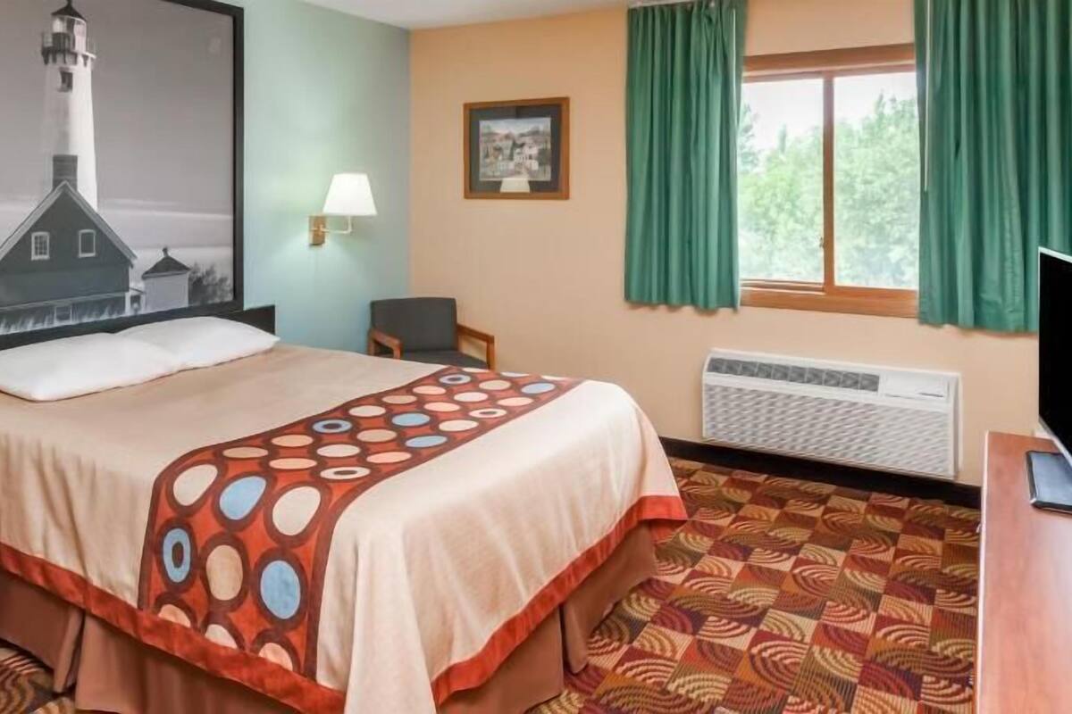 Habitación de hotel con cama, escritorio, silla, televisor, ventana con cortinas, radiador y un cuadro en la pared.
