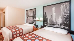A hotel room with two beds, each with a patterned bedspread, a nightstand with a lamp, and a framed cityscape picture on the wall. - Super 8 by Wyndham Shipshewana (Shipshewana)