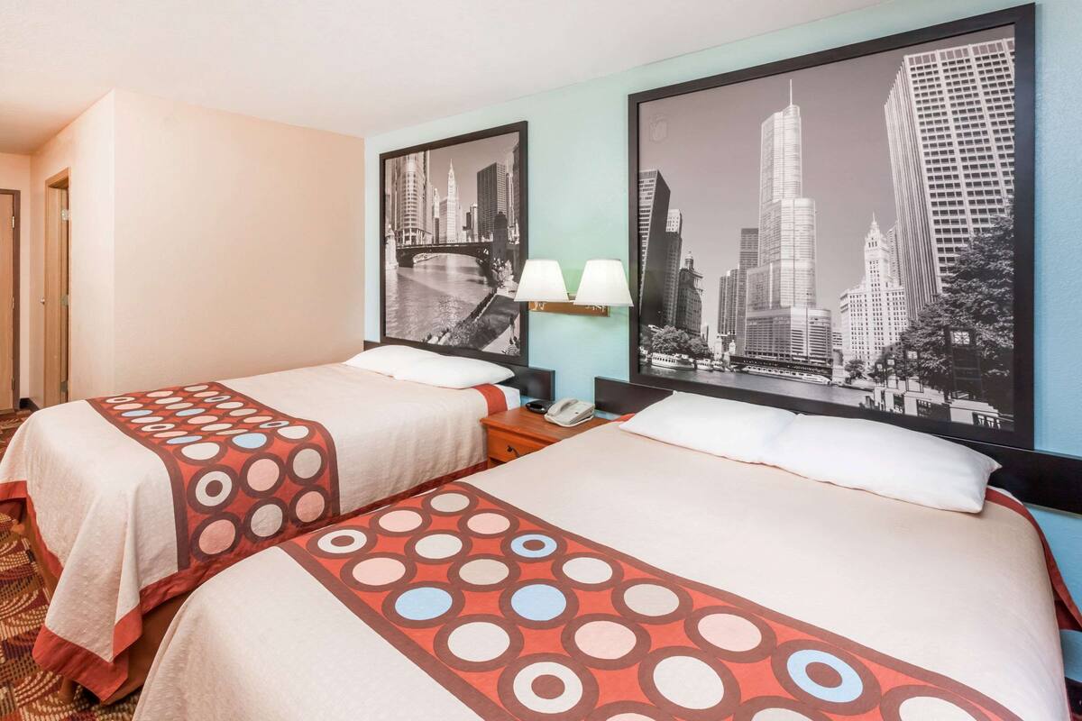 Una habitación de hotel con dos camas, cada una con un edredón estampado, una mesita de noche con lámpara y un cuadro con una vista urbana enmarcada en la pared.
