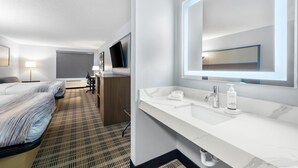 A hotel room with a bed, a desk, a chair, a desk lamp, a TV, a bathroom with a sink, mirror, and toiletries. - AmericInn by Wyndham Columbus Wisconsin (Columbus)