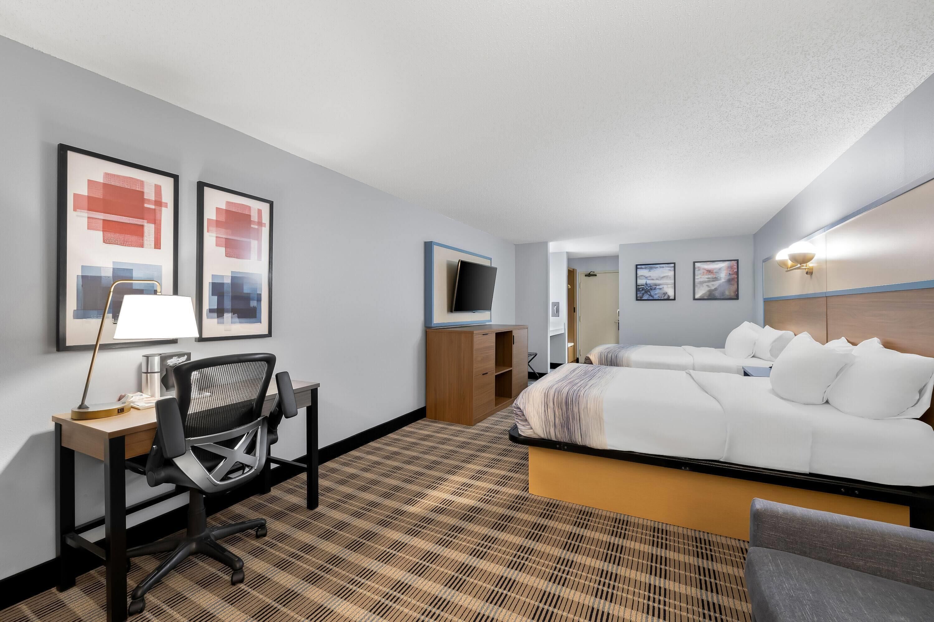 A hotel room with a bed, a desk with a chair, a TV, and artwork on the walls.