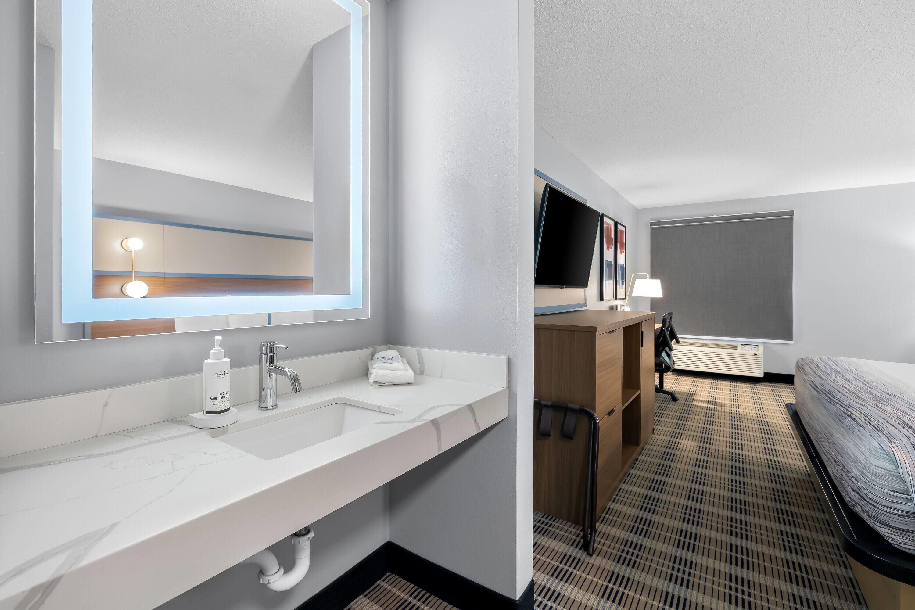 A modern hotel bathroom with a large mirror, white sink, and a towel dispenser.