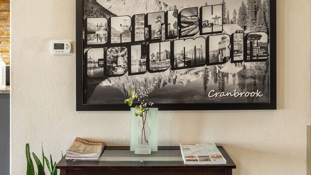 A framed welcome sign with a scenic landscape and the text "Greetings from Cranbrook" on a wall above a wooden console table with a vase of flowers and a magazine. - Ramada by Wyndham Cranbrook (Cranbrook)
