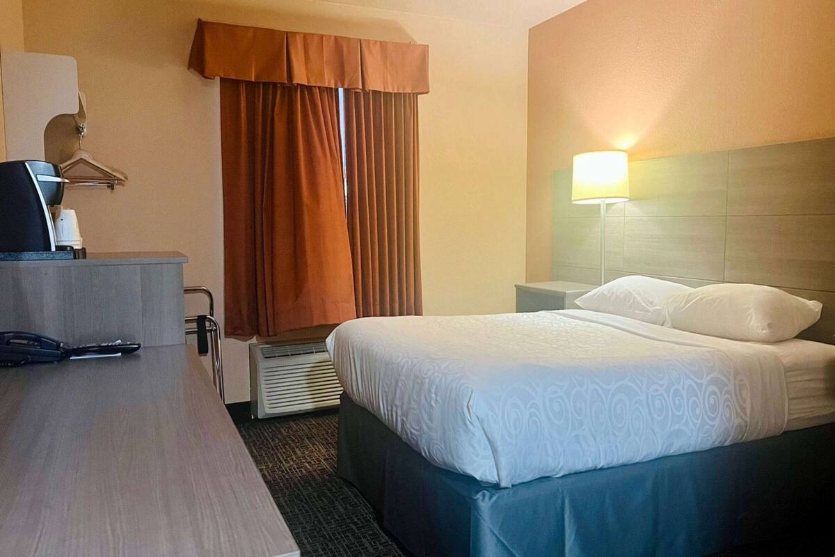 A hotel room with a bed, bedside table, lamp, and a desk with a computer and telephone.