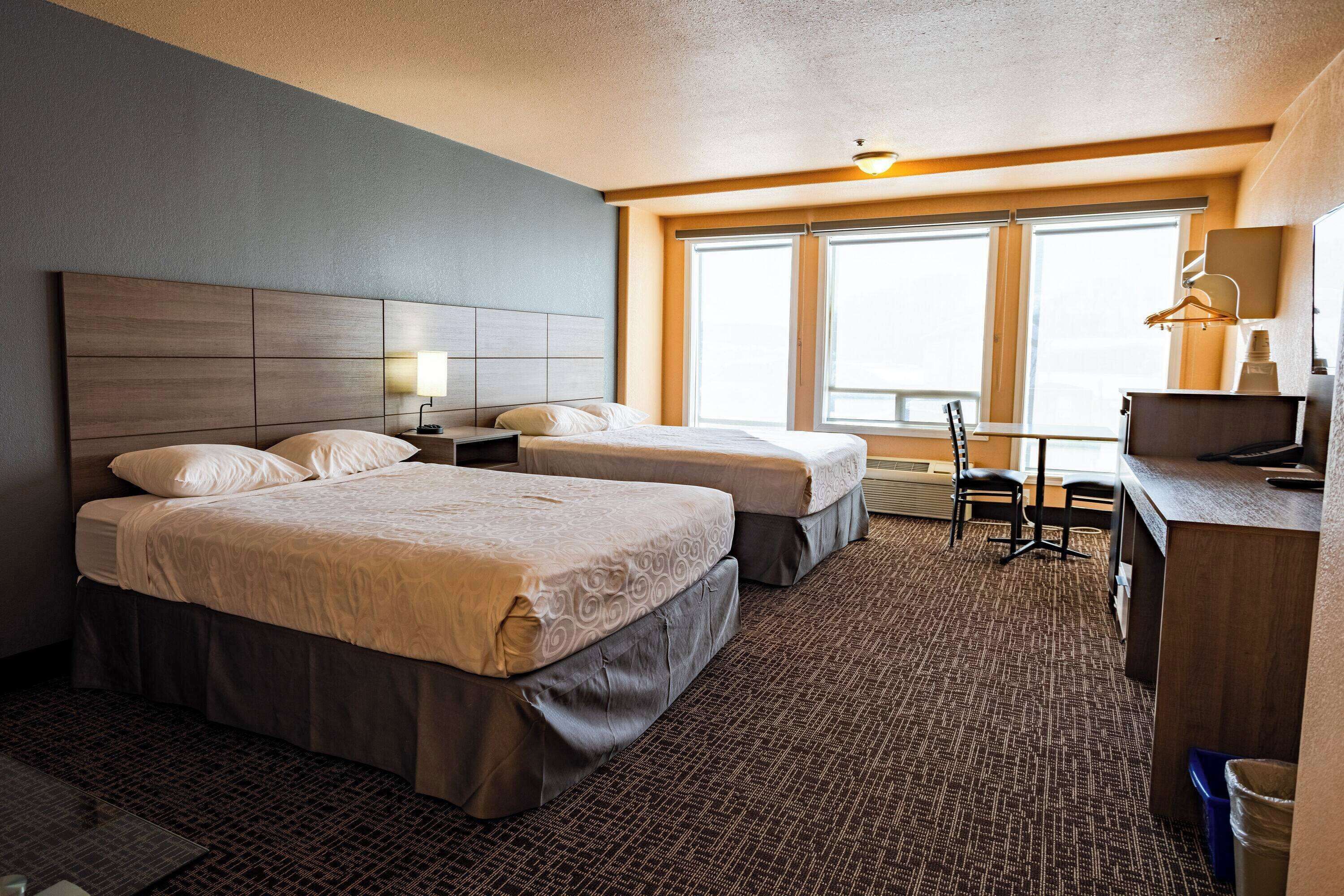 A hotel room with two beds, a desk, a chair, and a large window providing natural light.