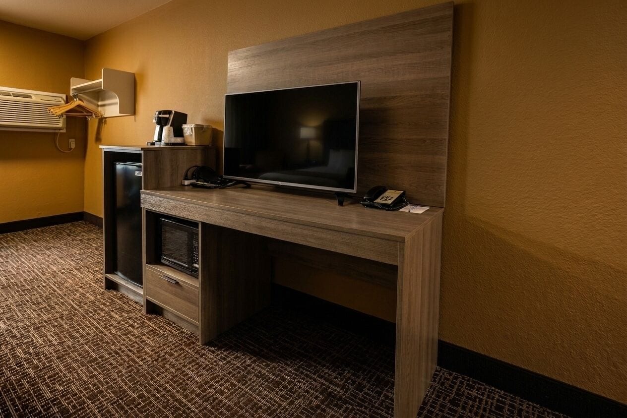 A hotel room with a wooden desk, a flat-screen TV, a coffee machine, a mini-fridge, and a microwave.