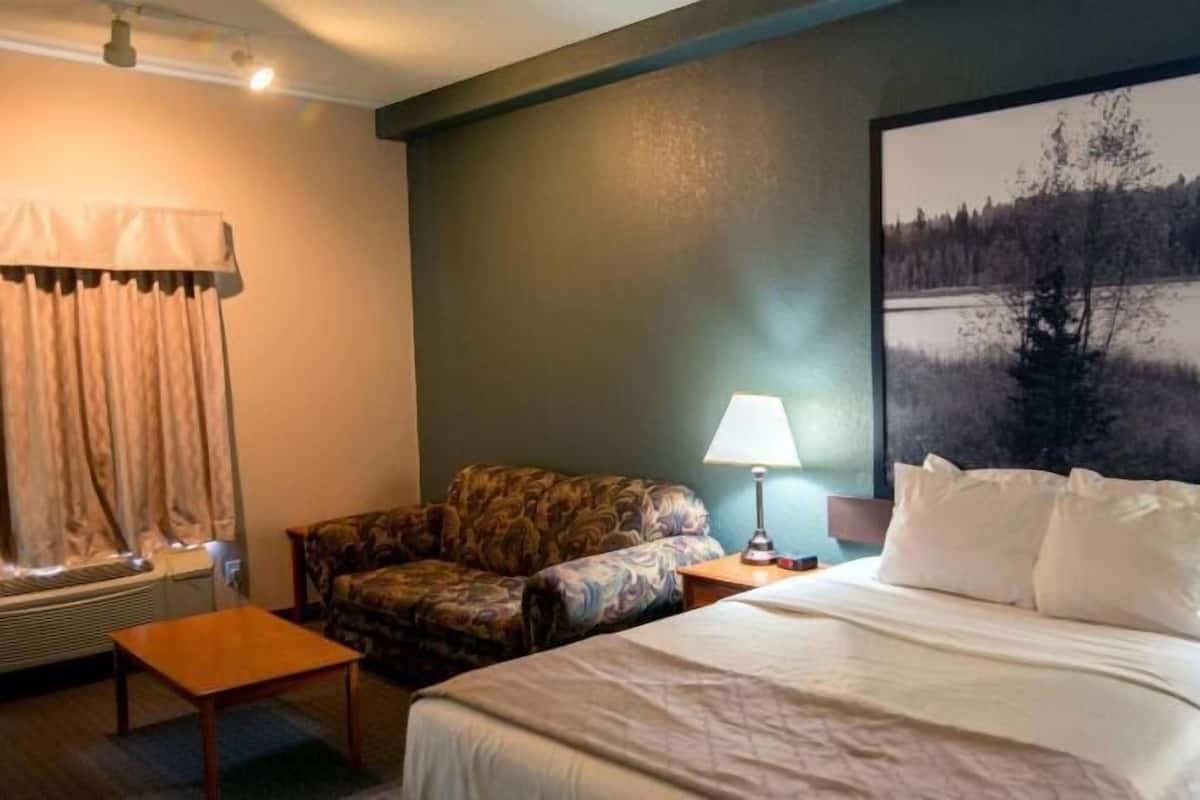 A hotel room with a bed, a chair, a table, a lamp, and a large painting of a forest scene.