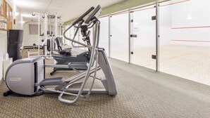 Ein Fitnessraum mit Cardiogeräten, einem an der Wand montierten Fernseher und einer Tür zu einem Squashplatz.