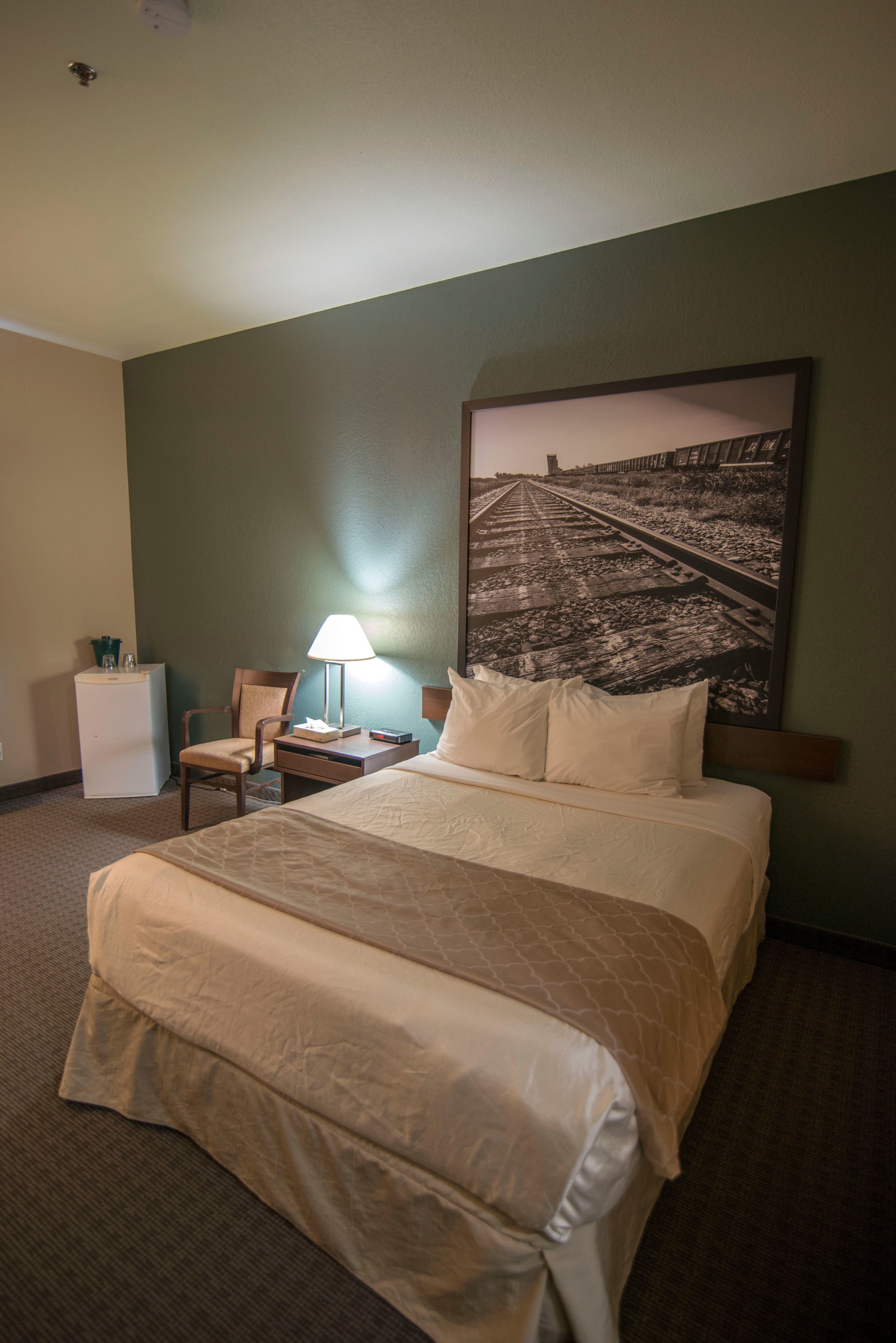 A hotel room with a large bed, a nightstand, a chair, a desk, a lamp, and a wall-mounted picture.