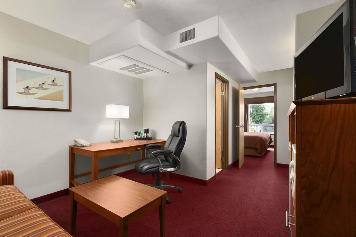 Ein Hotelzimmer mit einem Holztisch, einem schwarzen Bürostuhl, einer gestreiften Couch, einem Fernseher und einem Bett im Hintergrund.