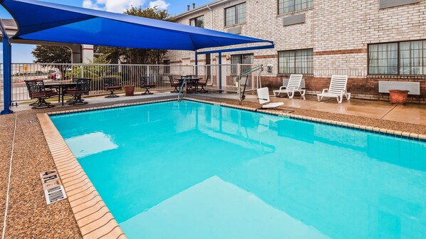 Una piscina con una cubierta azul, rodeada de sillones de descanso y un edificio de ladrillo al fondo.