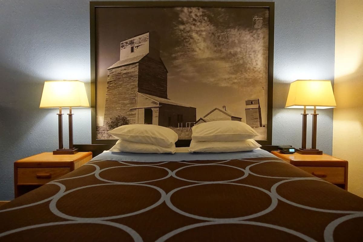 A hotel room with a bed, two bedside lamps, a large framed picture on the wall, and a patterned bedspread.