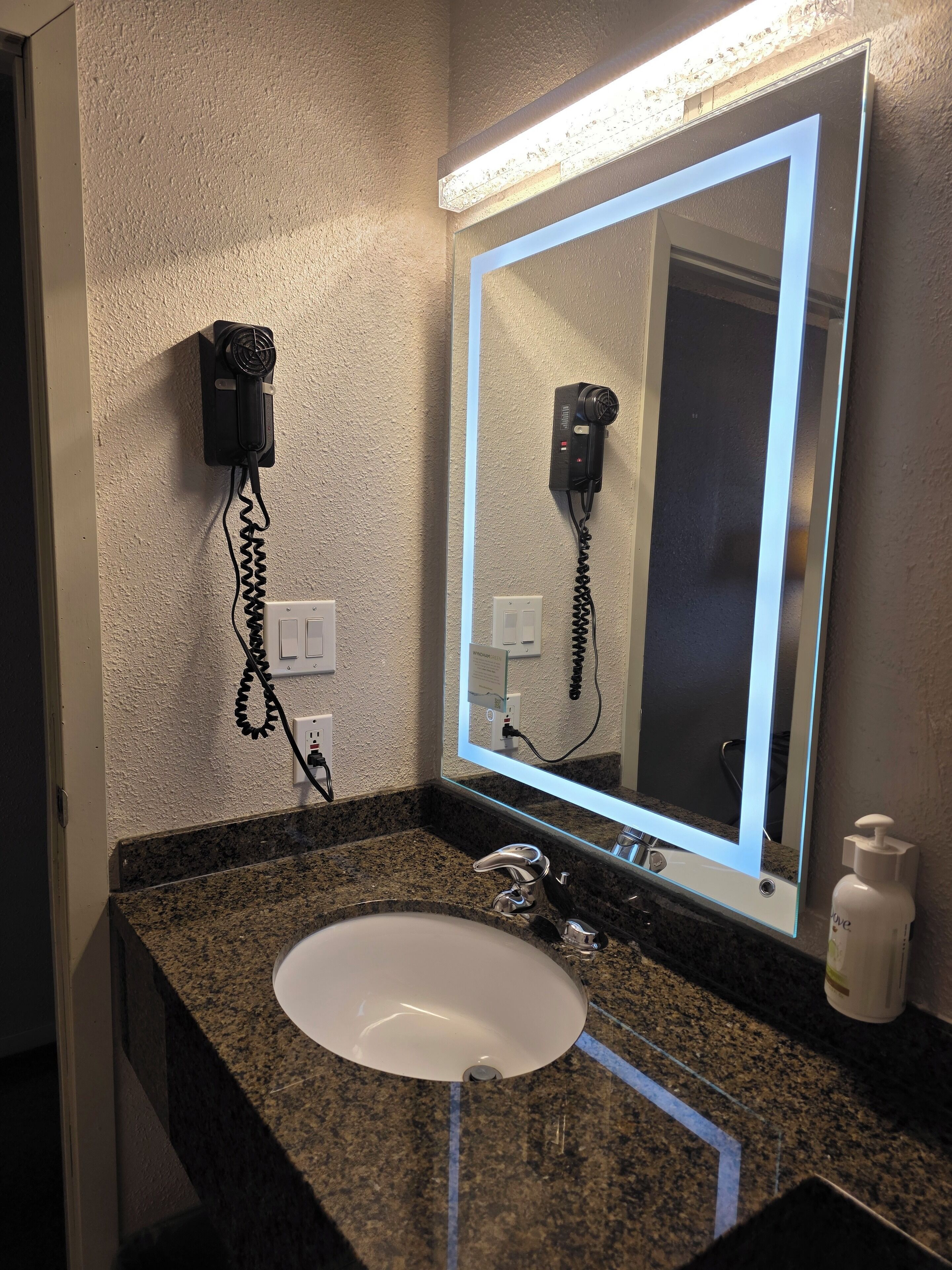 A bathroom with a granite countertop, a white sink, a mirror with a light, a hairdryer, and a phone.