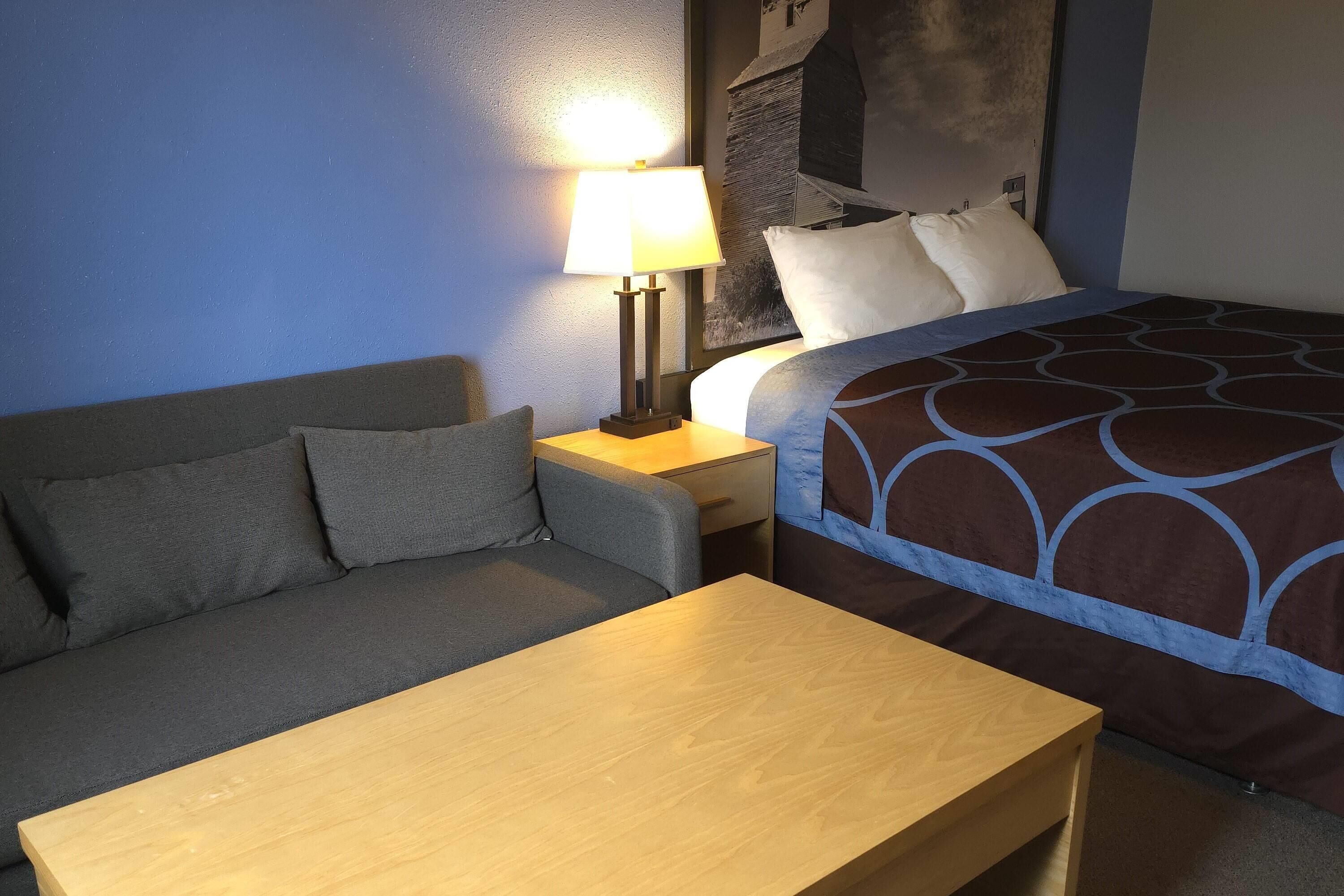 A hotel room with a bed, a sofa, a nightstand with a lamp, and a wooden table.