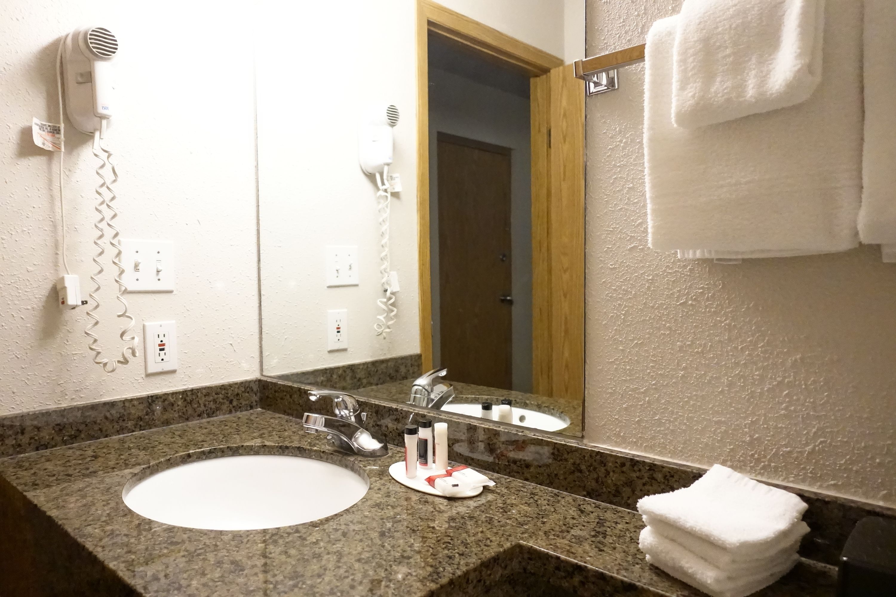 A hotel bathroom with a granite countertop sink, a mirror, a hairdryer, and a towel rack.