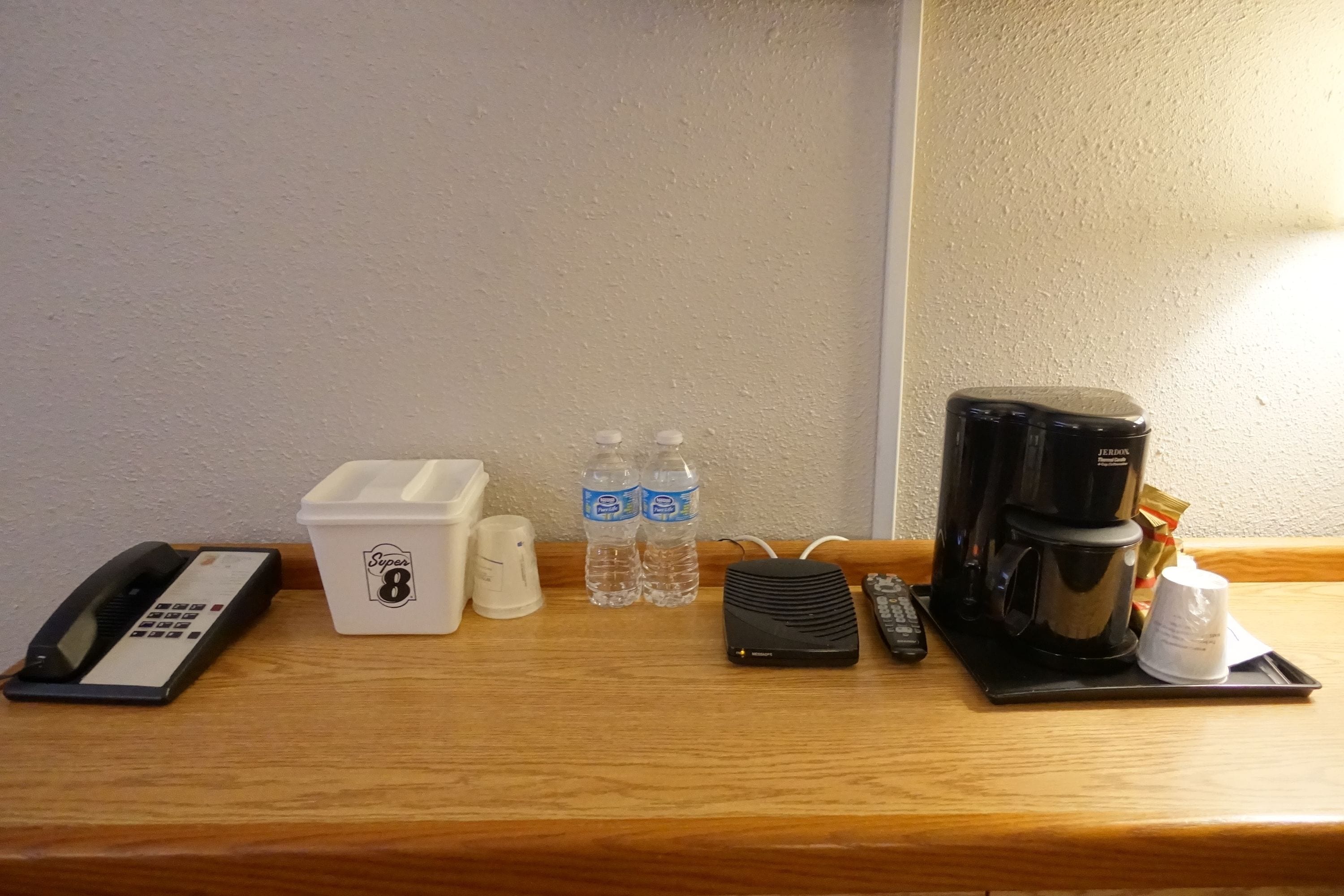 Zona de recepción de una habitación de hotel con un teléfono, un recipiente blanco con la etiqueta "Super 8", dos botellas de agua, un altavoz negro portátil, una cafetera y una taza de café sobre una bandeja.