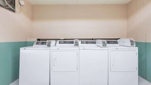 A row of four white front-loading washing machines in a laundry room with beige walls and teal accents.